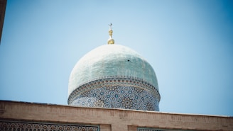 An ornate dome with intricate blue and white patterns against a clear blue sky. The dome features geometric and floral designs and is topped with a crescent moon symbol.