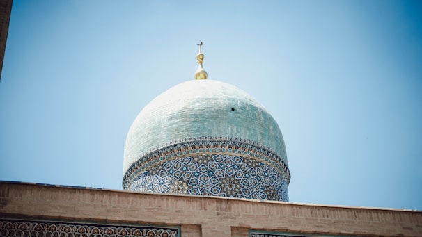 An ornate dome with intricate blue and white patterns against a clear blue sky. The dome features geometric and floral designs and is topped with a crescent moon symbol.