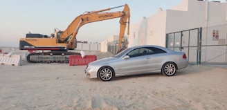 A silver coupe car is parked on a sandy area next to a construction site. An orange and black excavator with the brand name Hyundai is positioned nearby. Temporary barriers, some red and white, are placed around the site. In the background, white industrial buildings and a chain-link fence are visible under a clear sky.