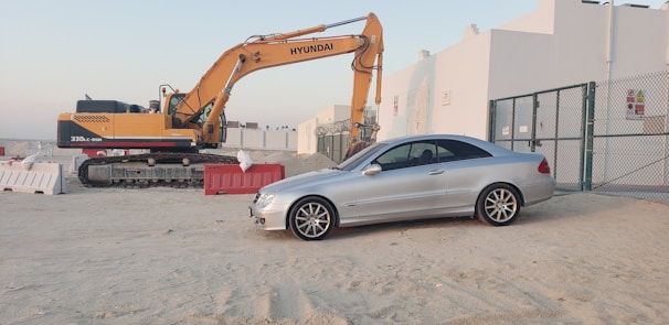 A silver coupe car is parked on a sandy area next to a construction site. An orange and black excavator with the brand name Hyundai is positioned nearby. Temporary barriers, some red and white, are placed around the site. In the background, white industrial buildings and a chain-link fence are visible under a clear sky.