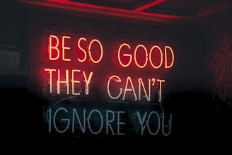 Neon signs display the motivational quote 'Be so good they can't ignore you' in a combination of red and white lights against a dark background.