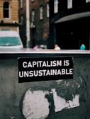 A sticker with white text on a black background reads 'Capitalism is Unsustainable.' It is placed on a weathered and possibly metallic surface with remnants of old stickers or paint. In the background, there is an urban street scene with blurred buildings and a van, suggesting a city setting.