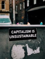 A sticker with white text on a black background reads 'Capitalism is Unsustainable.' It is placed on a weathered and possibly metallic surface with remnants of old stickers or paint. In the background, there is an urban street scene with blurred buildings and a van, suggesting a city setting.