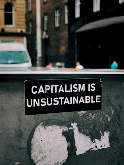 A sticker with white text on a black background reads 'Capitalism is Unsustainable.' It is placed on a weathered and possibly metallic surface with remnants of old stickers or paint. In the background, there is an urban street scene with blurred buildings and a van, suggesting a city setting.
