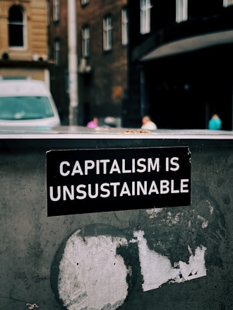 A sticker with white text on a black background reads 'Capitalism is Unsustainable.' It is placed on a weathered and possibly metallic surface with remnants of old stickers or paint. In the background, there is an urban street scene with blurred buildings and a van, suggesting a city setting.