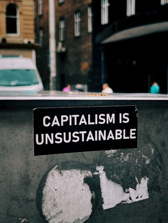 A sticker with white text on a black background reads 'Capitalism is Unsustainable.' It is placed on a weathered and possibly metallic surface with remnants of old stickers or paint. In the background, there is an urban street scene with blurred buildings and a van, suggesting a city setting.