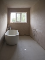 A minimalist bathroom with a freestanding bathtub positioned along one wall. The walls are unpainted, giving a raw, unfinished look. Large rectangular window at the far end brings natural light into the room. Plumbing fixtures appear bare, and an unconnected pipe is visible on the ground. The space is devoid of any decorative elements, emphasizing simplicity and functionality.