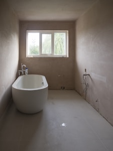 A minimalist bathroom with a freestanding bathtub positioned along one wall. The walls are unpainted, giving a raw, unfinished look. Large rectangular window at the far end brings natural light into the room. Plumbing fixtures appear bare, and an unconnected pipe is visible on the ground. The space is devoid of any decorative elements, emphasizing simplicity and functionality.