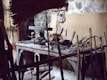 A rustic woodworking shop featuring a wooden workbench cluttered with various hand tools, such as saws, hammers, and chisels. The stone wall in the background provides an aged, historical feel. Wooden sticks and a woven bamboo panel provide a makeshift barricade at the forefront.