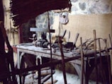A rustic woodworking shop featuring a wooden workbench cluttered with various hand tools, such as saws, hammers, and chisels. The stone wall in the background provides an aged, historical feel. Wooden sticks and a woven bamboo panel provide a makeshift barricade at the forefront.