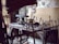 A rustic woodworking shop featuring a wooden workbench cluttered with various hand tools, such as saws, hammers, and chisels. The stone wall in the background provides an aged, historical feel. Wooden sticks and a woven bamboo panel provide a makeshift barricade at the forefront.