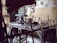 A rustic woodworking shop featuring a wooden workbench cluttered with various hand tools, such as saws, hammers, and chisels. The stone wall in the background provides an aged, historical feel. Wooden sticks and a woven bamboo panel provide a makeshift barricade at the forefront.