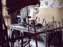 A rustic woodworking shop featuring a wooden workbench cluttered with various hand tools, such as saws, hammers, and chisels. The stone wall in the background provides an aged, historical feel. Wooden sticks and a woven bamboo panel provide a makeshift barricade at the forefront.
