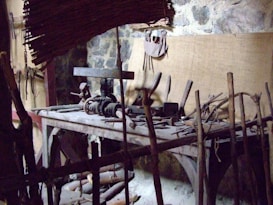 A rustic woodworking shop featuring a wooden workbench cluttered with various hand tools, such as saws, hammers, and chisels. The stone wall in the background provides an aged, historical feel. Wooden sticks and a woven bamboo panel provide a makeshift barricade at the forefront.