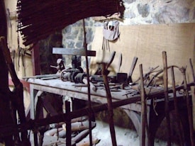 A rustic woodworking shop featuring a wooden workbench cluttered with various hand tools, such as saws, hammers, and chisels. The stone wall in the background provides an aged, historical feel. Wooden sticks and a woven bamboo panel provide a makeshift barricade at the forefront.