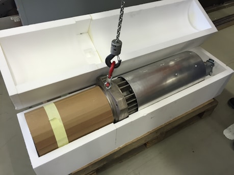 A large metal cylindrical object is partially placed inside a custom-fitted white foam case. The cylinder has a cardboard covering on one end and is lifted by a metal hook attached to a chain. It appears to be on a wooden pallet.