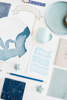 A flat lay of various items including a decorative plate with an abstract blue design, a pale blue mug, a blue donut on a plate, and a card saying 'Hip Hip Hooray!'. Surrounding the main items are small star-shaped decorations, fabric swatches, a pencil, and a decorative banner with the phrase 'Best day ever'.