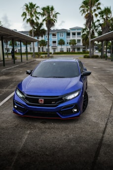 A sleek, blue Honda car parked in an outdoor area with wet pavement, surrounded by palm trees and residential buildings in the background on a cloudy day.