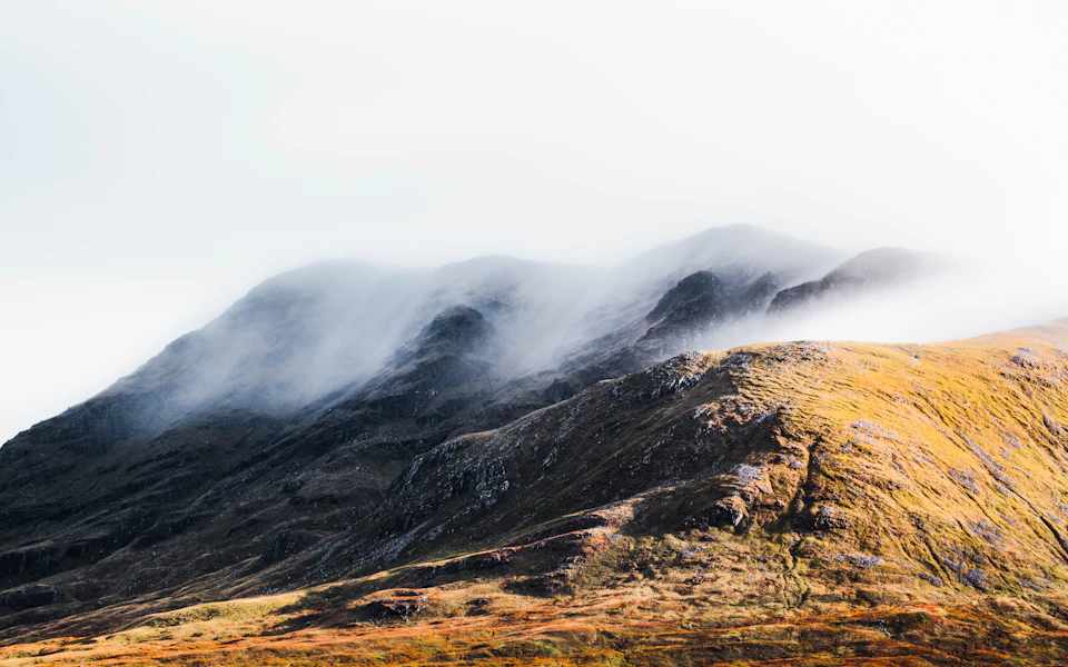 The image depicts a majestic mountain range with a hazy, foggy atmosphere. The mountains are covered in a layer of grayish-brown fog that obscures their details and adds an ethereal quality to the scene. The perspective is from below, looking up at the towering peaks against a light brown sky. The colors in the image are predominantly gray and brown, with hints of green and yellow visible on the mountainside.