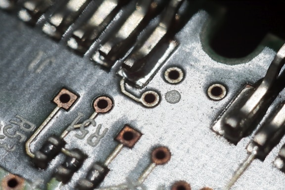 A close-up view of a circuit board showing metallic tracks, solder points, and various electronic components. The image highlights the intricate design and small details of the board with a combination of silver, copper, and greenish tones.