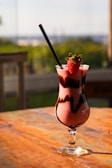 A glass of strawberry smoothie sits on a wooden table. The smoothie is decorated with chocolate swirls, and a fresh strawberry is placed on the rim of the glass. Sunlight illuminates the drink, creating a warm, inviting ambiance. A black straw is placed in the glass. The background is softly blurred, hinting at an outdoor setting with greenery.