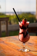 A glass of strawberry smoothie sits on a wooden table. The smoothie is decorated with chocolate swirls, and a fresh strawberry is placed on the rim of the glass. Sunlight illuminates the drink, creating a warm, inviting ambiance. A black straw is placed in the glass. The background is softly blurred, hinting at an outdoor setting with greenery.