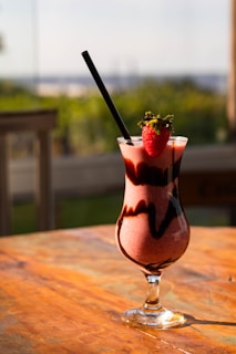 A glass of strawberry smoothie sits on a wooden table. The smoothie is decorated with chocolate swirls, and a fresh strawberry is placed on the rim of the glass. Sunlight illuminates the drink, creating a warm, inviting ambiance. A black straw is placed in the glass. The background is softly blurred, hinting at an outdoor setting with greenery.