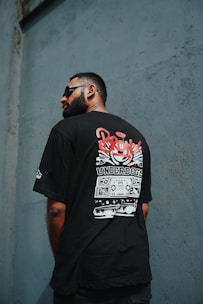 A man with a beard and sunglasses is facing away, displaying a T-shirt with a graphic design featuring the word 'Bronx' and 'Underdogs'. The background consists of a textured wall in a shade of blue-gray.