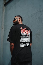A man with a beard and sunglasses is facing away, displaying a T-shirt with a graphic design featuring the word 'Bronx' and 'Underdogs'. The background consists of a textured wall in a shade of blue-gray.