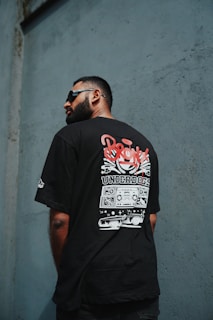 A man with a beard and sunglasses is facing away, displaying a T-shirt with a graphic design featuring the word 'Bronx' and 'Underdogs'. The background consists of a textured wall in a shade of blue-gray.
