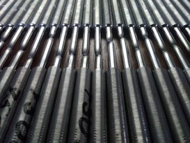 A close-up view of a series of metallic rods neatly arranged in parallel rows. The rods have a shiny, reflective surface that catches the light, and they're stamped with text near the ends.