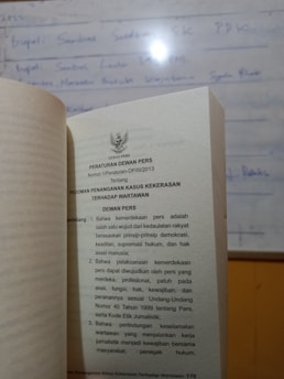 A book is open to a page containing text in Indonesian about press regulations, with a blurred whiteboard in the background. The text discusses issues concerning press freedom and the safety of journalists.