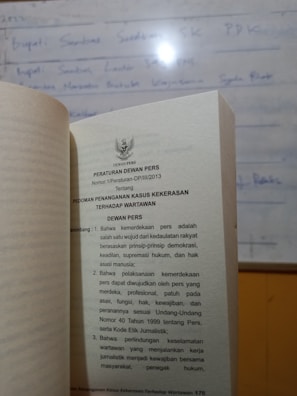 A book is open to a page containing text in Indonesian about press regulations, with a blurred whiteboard in the background. The text discusses issues concerning press freedom and the safety of journalists.