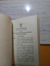 A book is open to a page containing text in Indonesian about press regulations, with a blurred whiteboard in the background. The text discusses issues concerning press freedom and the safety of journalists.
