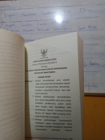 A book is open to a page containing text in Indonesian about press regulations, with a blurred whiteboard in the background. The text discusses issues concerning press freedom and the safety of journalists.