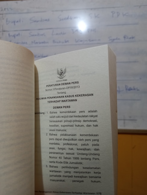 A book is open to a page containing text in Indonesian about press regulations, with a blurred whiteboard in the background. The text discusses issues concerning press freedom and the safety of journalists.