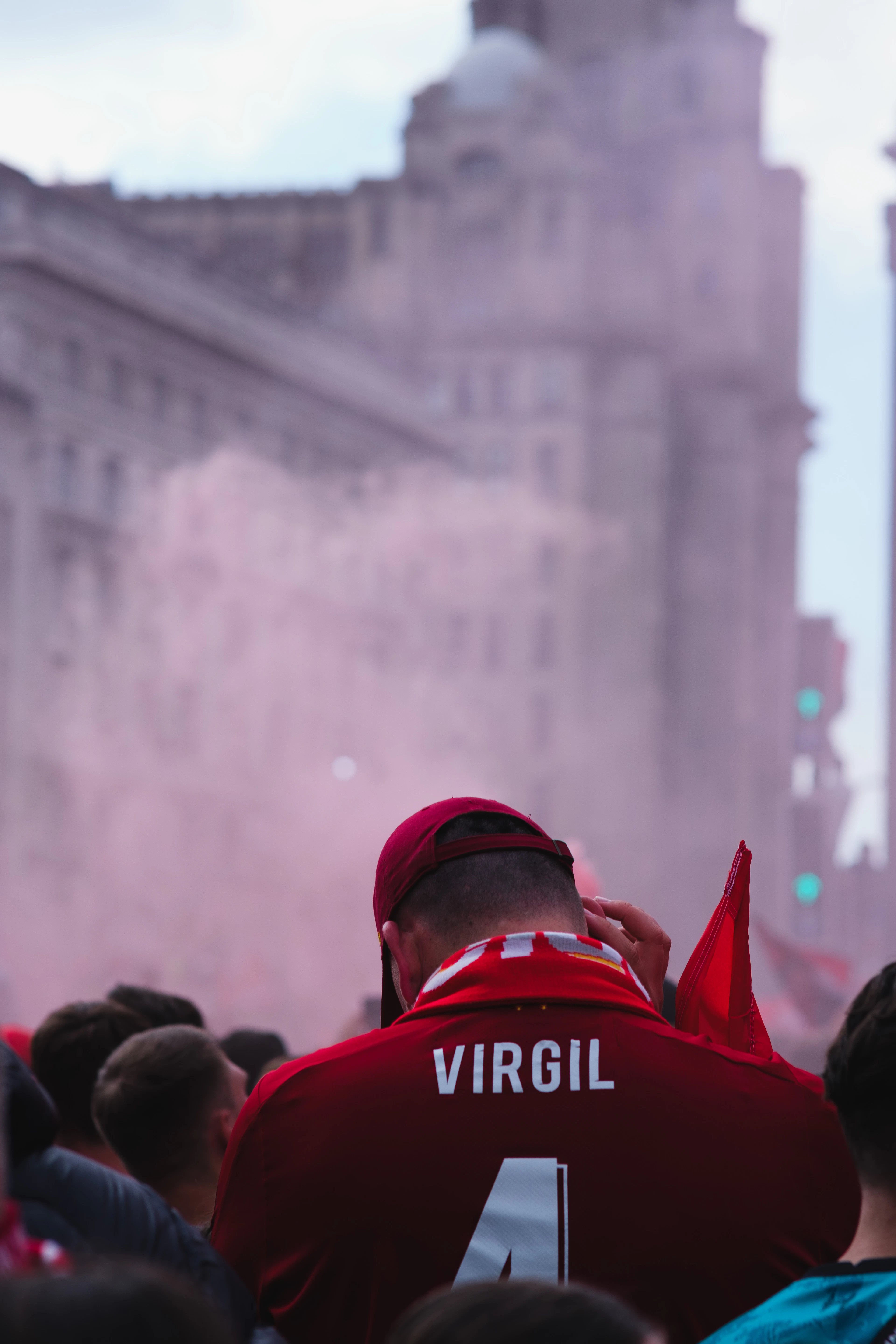 A football fan in a 'Virgil' shirt amid pink smoke flares during a trophy parade, waterfront buildings blurred behind.
