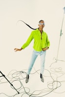 A person wearing a bright green button-up shirt and light blue jeans stands confidently in a studio setting. They have a distinctive long braided hairstyle and are accessorized with white, futuristic sunglasses and black boots with pointed toes. The studio setup includes visible lighting equipment and cables scattered on the floor, creating an artistic and dynamic composition.