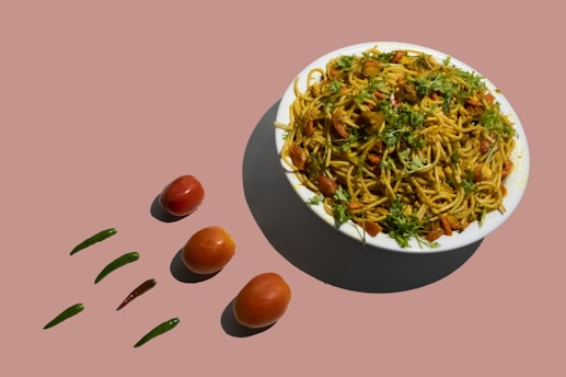 A bowl of noodles garnished with chopped herbs, surrounded by whole tomatoes and green chili peppers arranged on a solid colored surface.