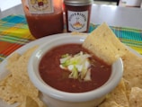 A bowl of salsa topped with sour cream and chopped lettuce is surrounded by tortilla chips. A larger jar of salsa and a smaller jar with a spicy label are in the background. A colorful woven cloth adds a vibrant touch to the scene.