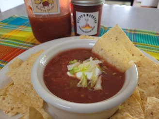 A bowl of salsa topped with sour cream and chopped lettuce is surrounded by tortilla chips. A larger jar of salsa and a smaller jar with a spicy label are in the background. A colorful woven cloth adds a vibrant touch to the scene.