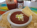 A bowl of salsa topped with sour cream and chopped lettuce is surrounded by tortilla chips. A larger jar of salsa and a smaller jar with a spicy label are in the background. A colorful woven cloth adds a vibrant touch to the scene.