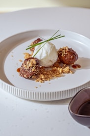 A white plate with three brown, round fried desserts garnished with crumbs and sauce. In the center, there is a mound of whipped cream or ice cream, topped with two green chive stalks. A small brown sauce dish is placed next to the plate.