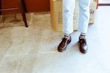 A pair of feet wearing black shoes with patterned black and white socks, standing on a cracked concrete floor. The person is wearing light blue jeans. In the background, a large cardboard box with Asian characters is partially visible.