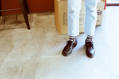 A pair of feet wearing black shoes with patterned black and white socks, standing on a cracked concrete floor. The person is wearing light blue jeans. In the background, a large cardboard box with Asian characters is partially visible.