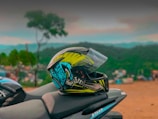 A vibrant motorcycle helmet with graphic designs sits on the seat of a motorcycle against a blurred scenic background featuring hills and vegetation. The helmet has a mix of yellow, blue, and black colors.