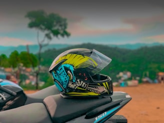 A vibrant motorcycle helmet with graphic designs sits on the seat of a motorcycle against a blurred scenic background featuring hills and vegetation. The helmet has a mix of yellow, blue, and black colors.