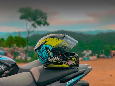 A vibrant motorcycle helmet with graphic designs sits on the seat of a motorcycle against a blurred scenic background featuring hills and vegetation. The helmet has a mix of yellow, blue, and black colors.