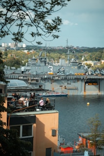 A large military ship is docked in a harbor, surrounded by water and greenery in the background. In the foreground, people are sitting on the rooftop terrace of a nearby building enjoying the view. The scene captures a blend of urban life and military presence.