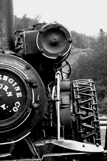 A close-up view of a vintage steam locomotive, focusing on its intricate mechanical components and weathered metal surfaces. The image highlights the round, heavy metal structures and the faded text on the side of the engine. A backdrop of trees and a misty sky adds a sense of nostalgia.